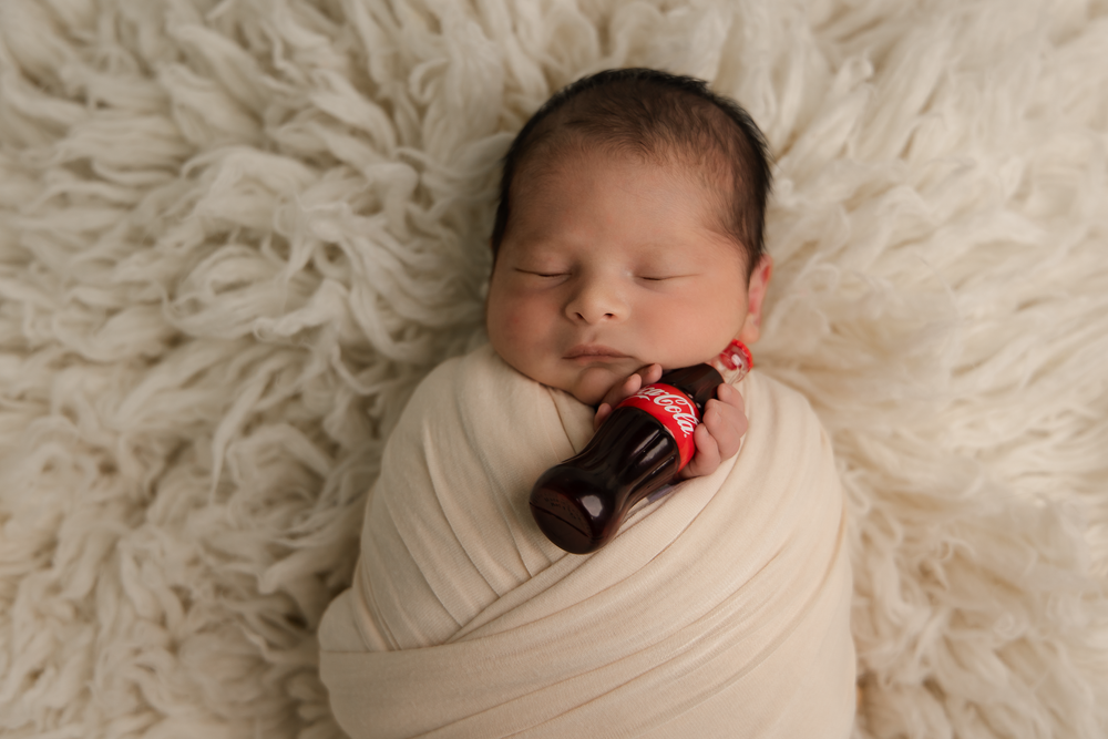 Swaddled newborn baby sleeping on fluffy white rug holding mini Coca-Cola bottle.
