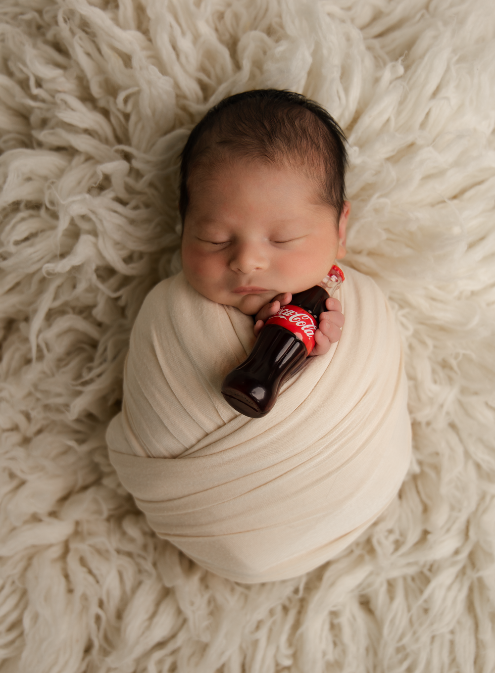 Newborn baby swaddled in cream wrap sleeping on fluffy white rug holding a miniature Coca-Cola bottle.