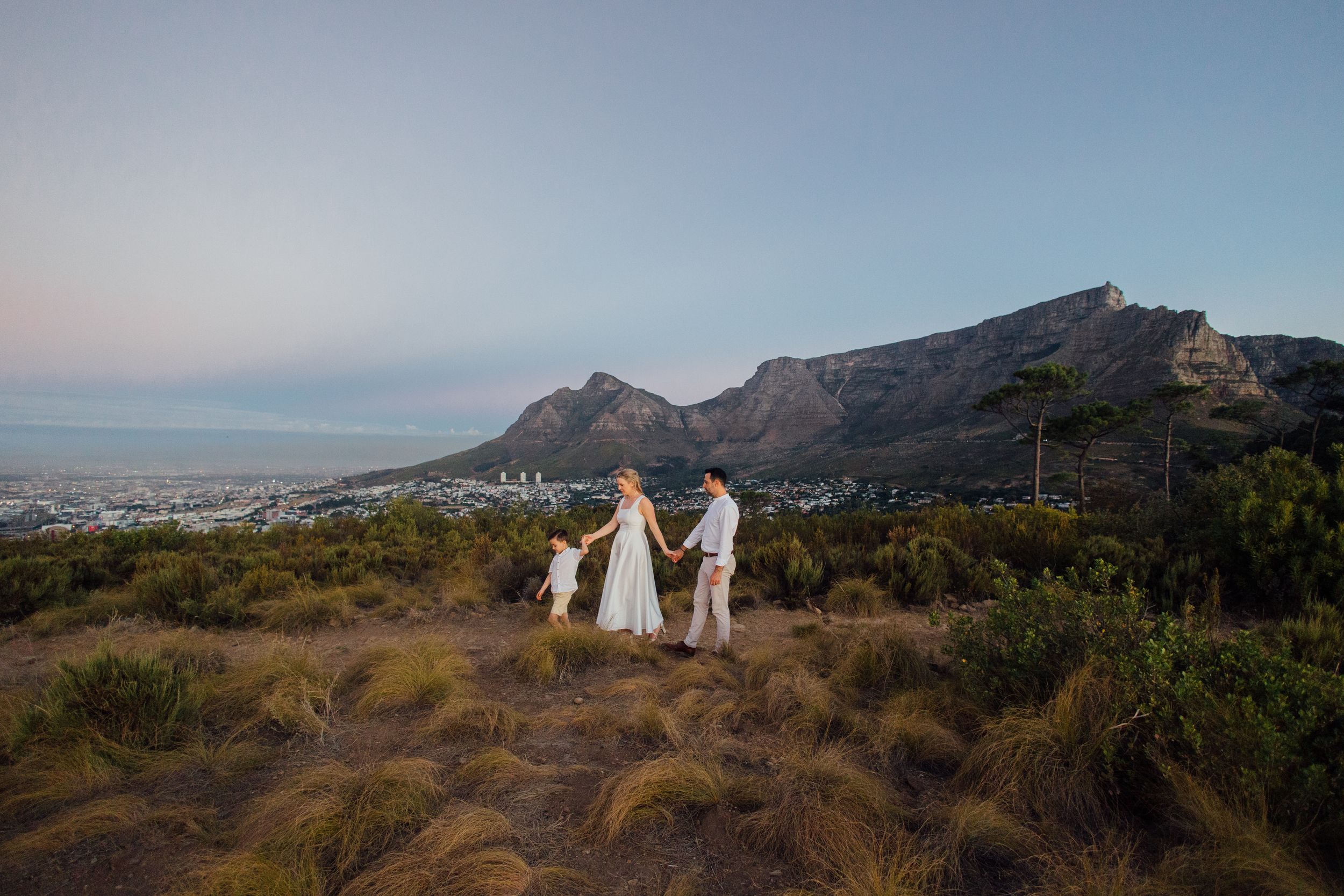 Family in white holding hands on a scenic Cape Town hillside with Table Mountain backdrop at dusk.