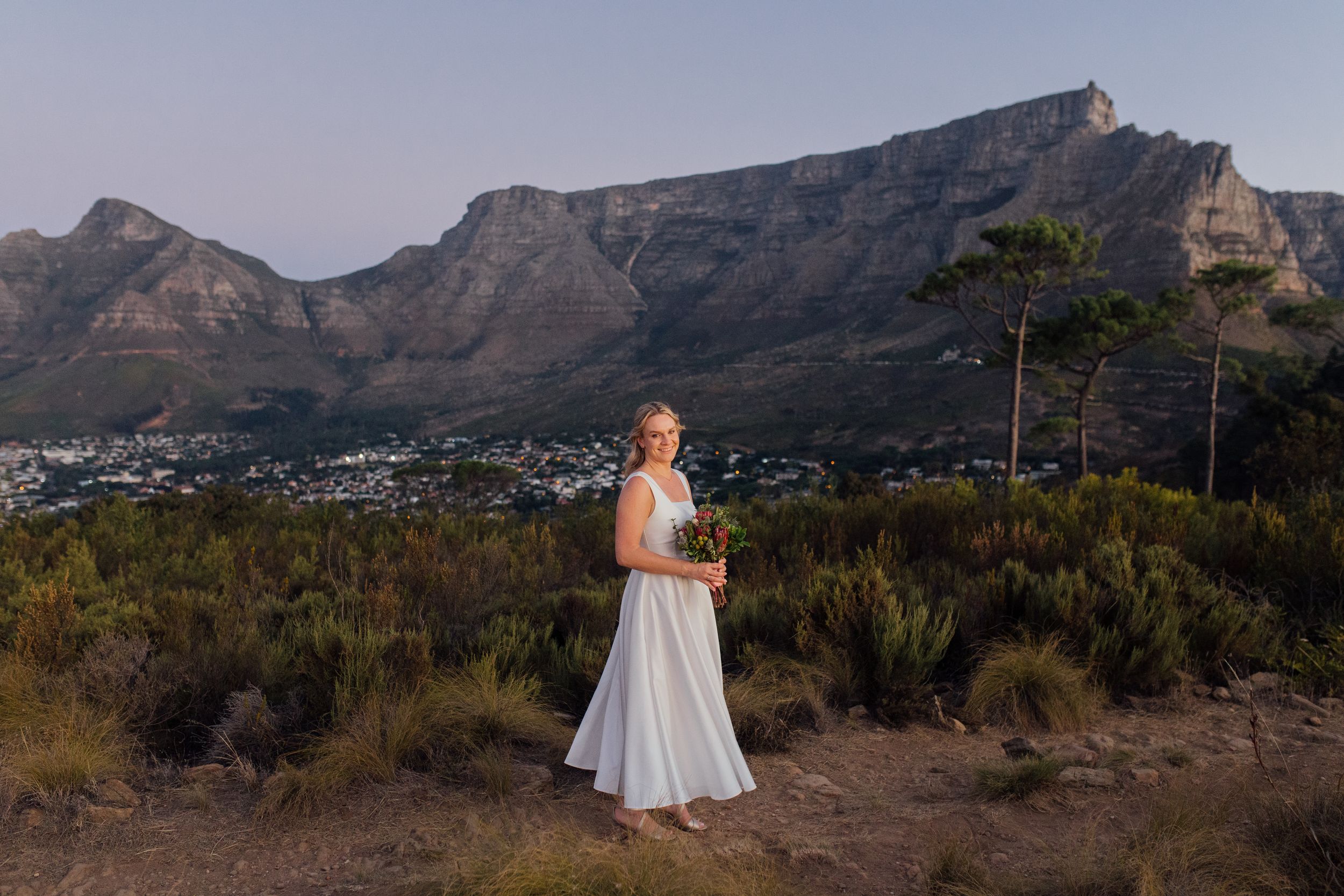 Bride in white gown holding bouquet with Table Mountain and Cape Town cityscape backdrop at sunset.