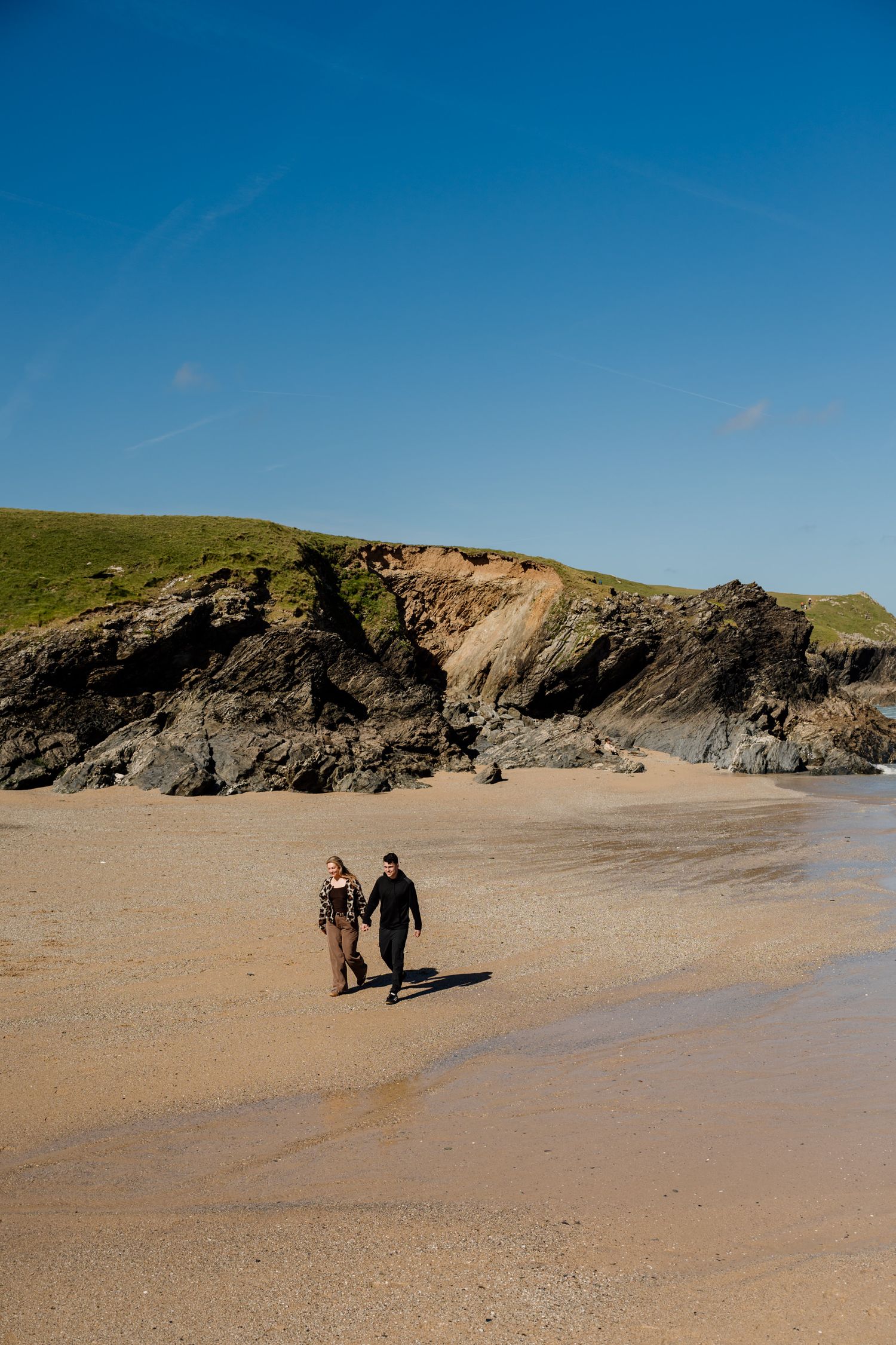 Couple walking hand in hand on a sandy beach with rocky cliffs and green hills under a clear blue sky.