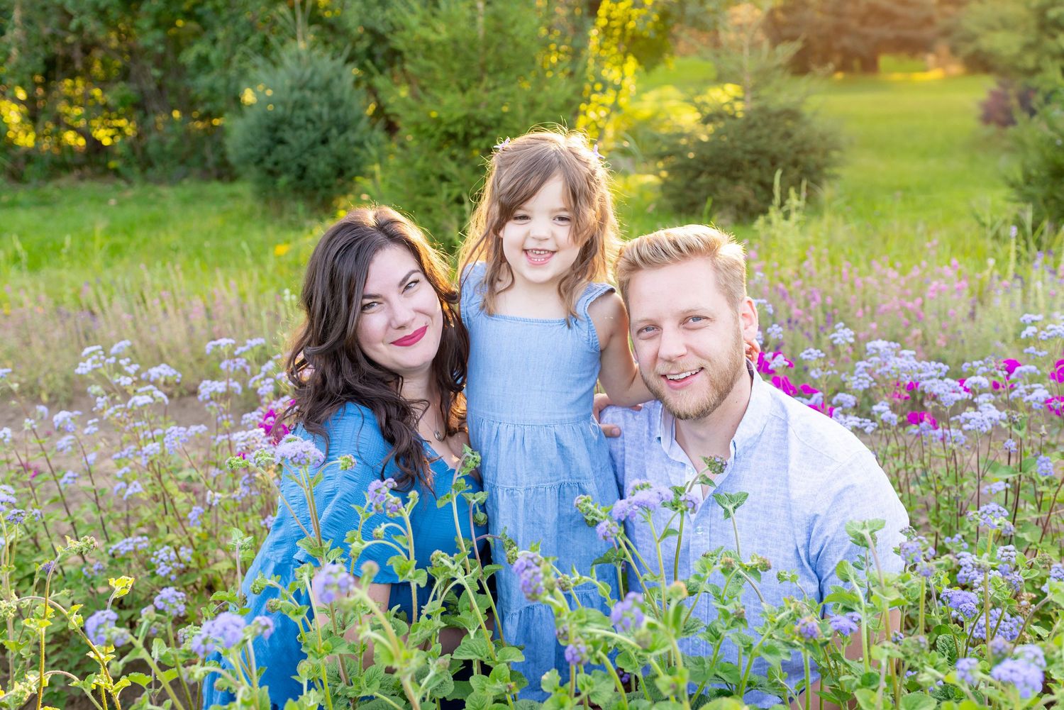 Family of three smiling in a sunny wildflower field, mom and dad seated with young daughter between them.