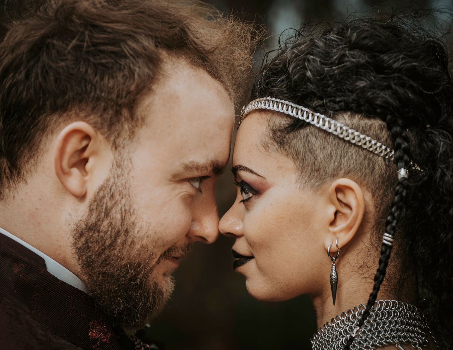 A couple with bohemian style facing each other closely, woman with shaved sides and chain headband, man with beard.