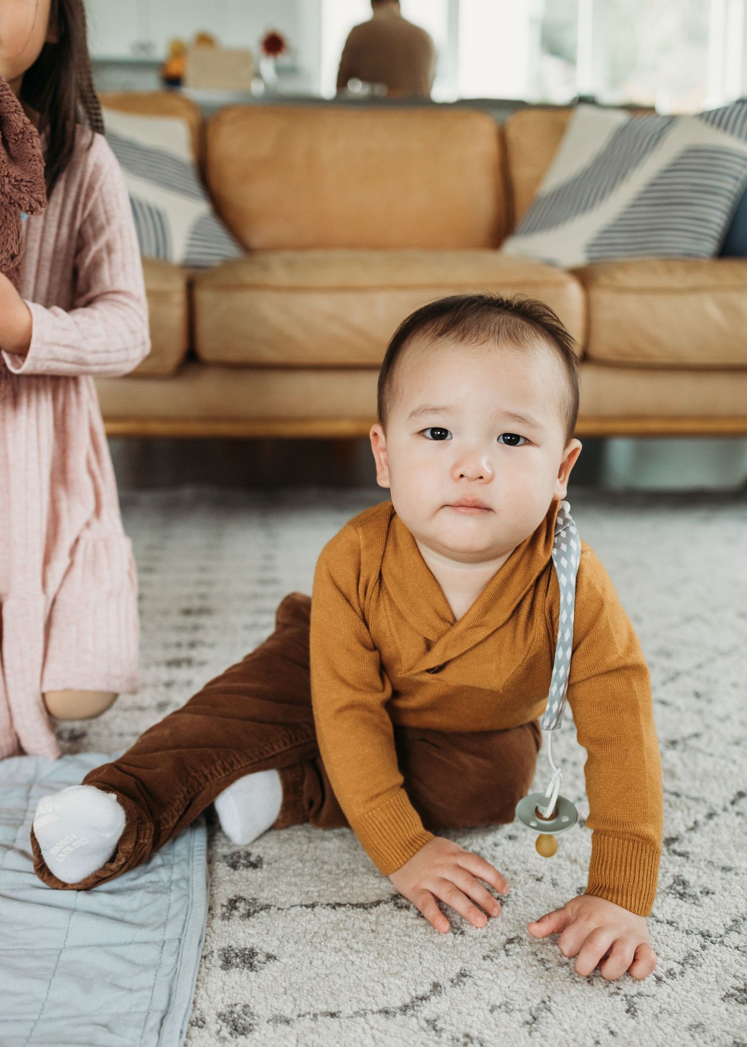 Baby in mustard yellow cardigan crawls on patterned carpet in living room setting.