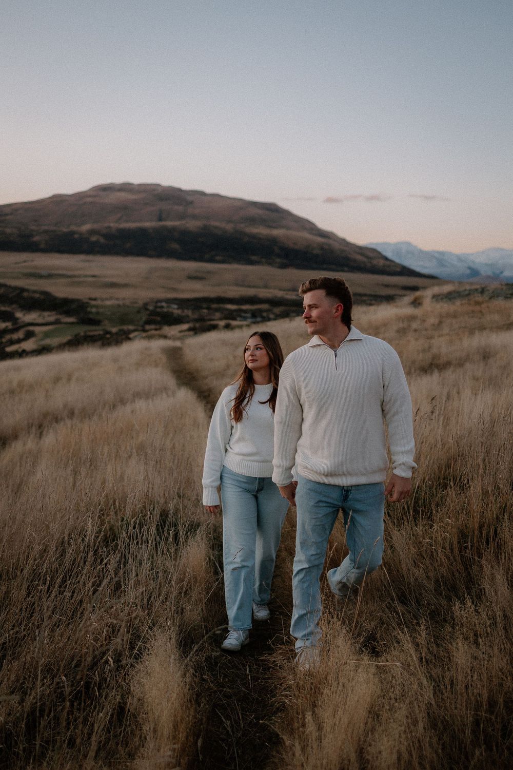 Couple in white sweaters and jeans walking through golden grass fields with mountain backdrop at dusk.
