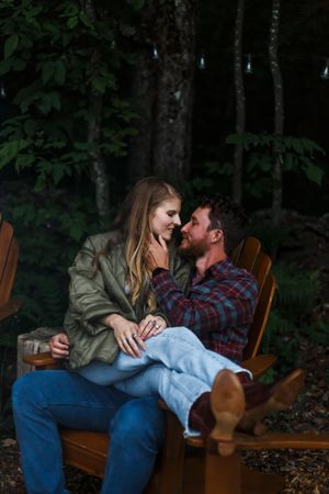 A couple shares an intimate moment while relaxing on wooden chairs in a wooded setting.