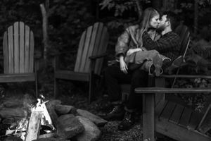 A black and white photo of a couple by a campfire with Adirondack chairs.