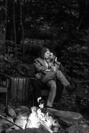 A couple cuddles on Adirondack chairs next to a glowing campfire in a dark forest setting.