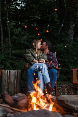 A couple sits close together by a glowing campfire with string lights twinkling in the forest background.