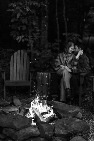 A couple sits close together on outdoor chairs with a blazing bonfire in the foreground.