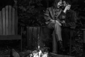 A group gathers around a glowing campfire at night with Adirondack chairs visible.