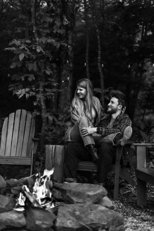 A couple relaxes in Adirondack chairs beside a crackling campfire in the woods.