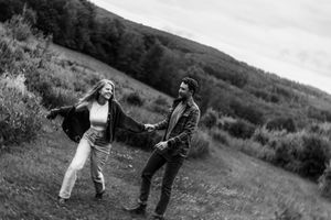 A couple dances playfully in a grassy meadow with rolling hills in the background on an overcast day.
