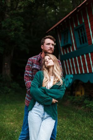 A couple embraces near a vintage red and turquoise camper trailer surrounded by trees.