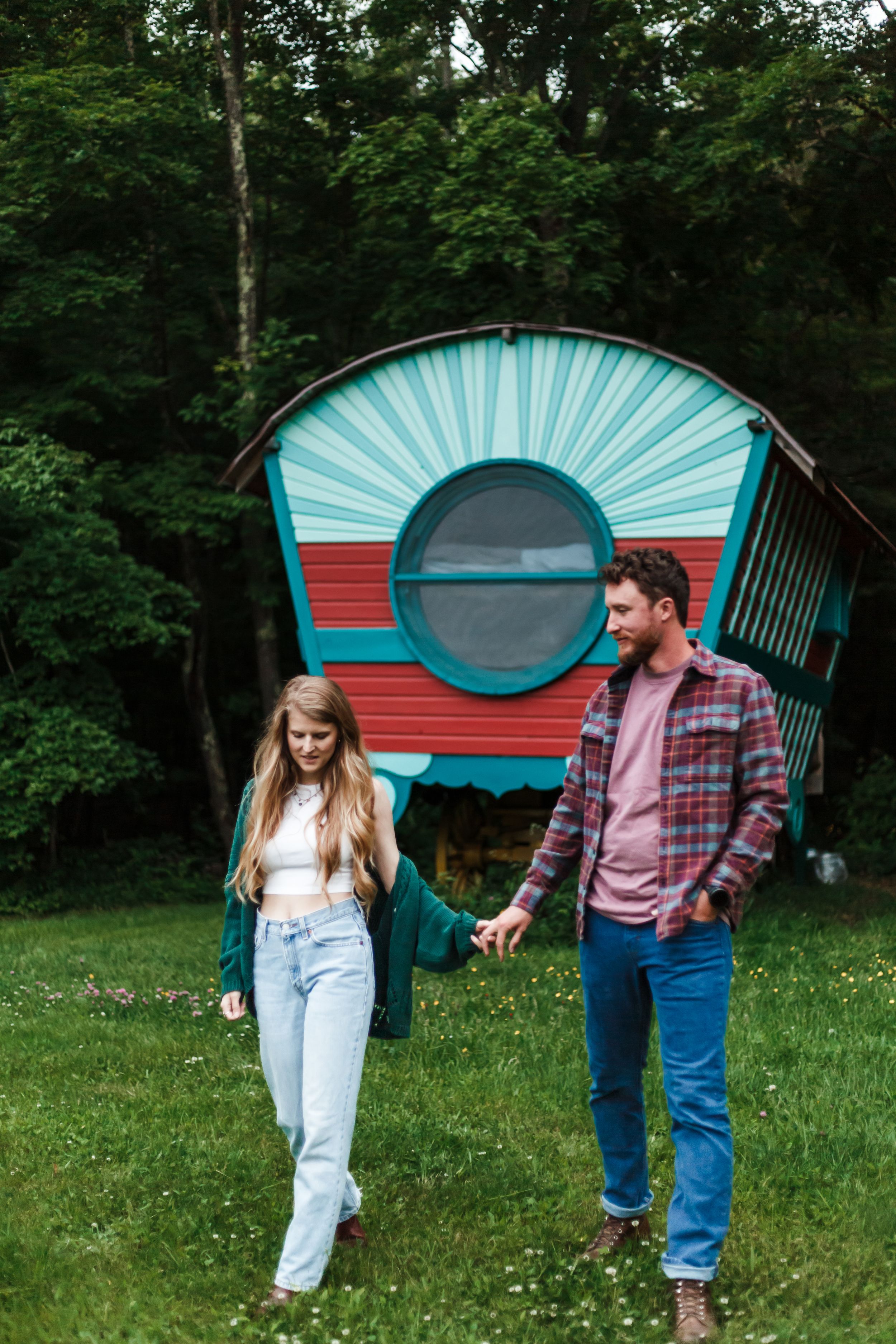A couple dances playfully in front of a red and turquoise vintage camper trailer in a wooded setting.