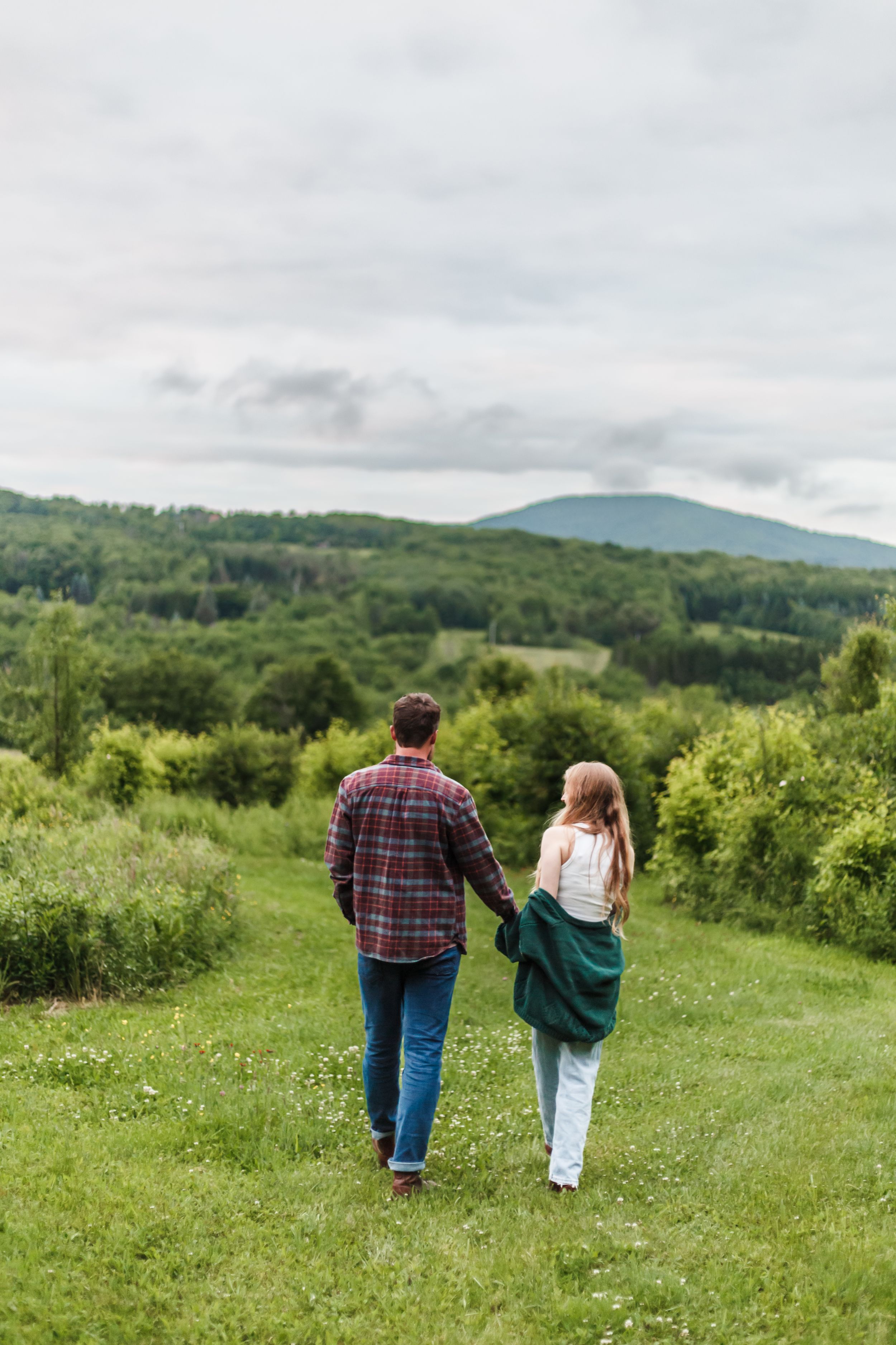 A couple walks hand in hand through a lush green meadow with mountains in the distance on a cloudy day.