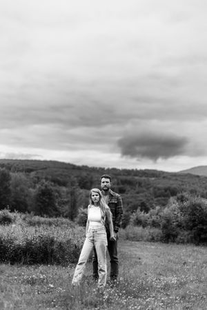 Two people stand together in a wildflower field against a backdrop of lush green hills and cloudy skies.