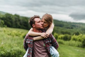 Two people embrace and kiss while standing in a lush green mountainside meadow.