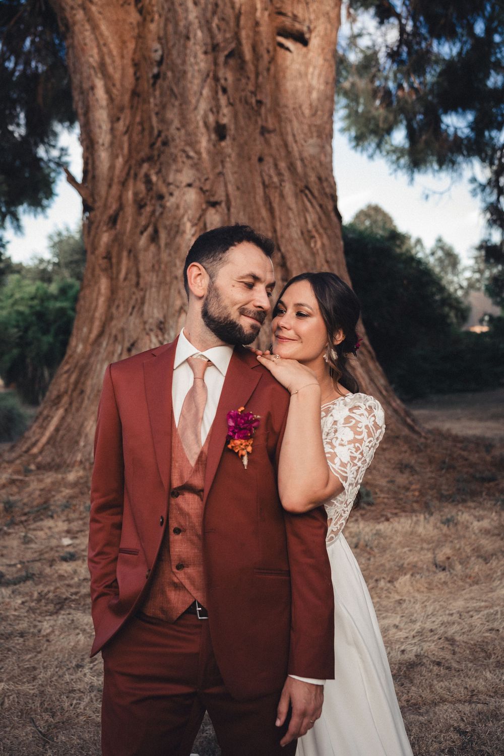 Bride in white lace dress leans on groom in burgundy suit with floral boutonniere, posing before a large redwood tree.