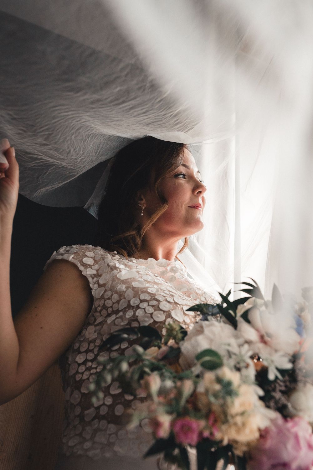 Bride in lace gown holds bouquet under flowing veil by window, gazing upward in soft natural light.