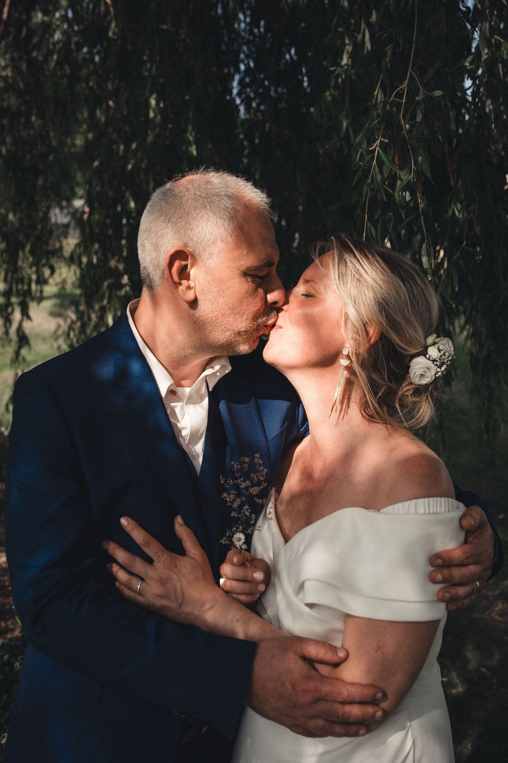 A couple shares a romantic kiss under a weeping willow tree, the bride in an off-shoulder white gown and groom in a navy suit.