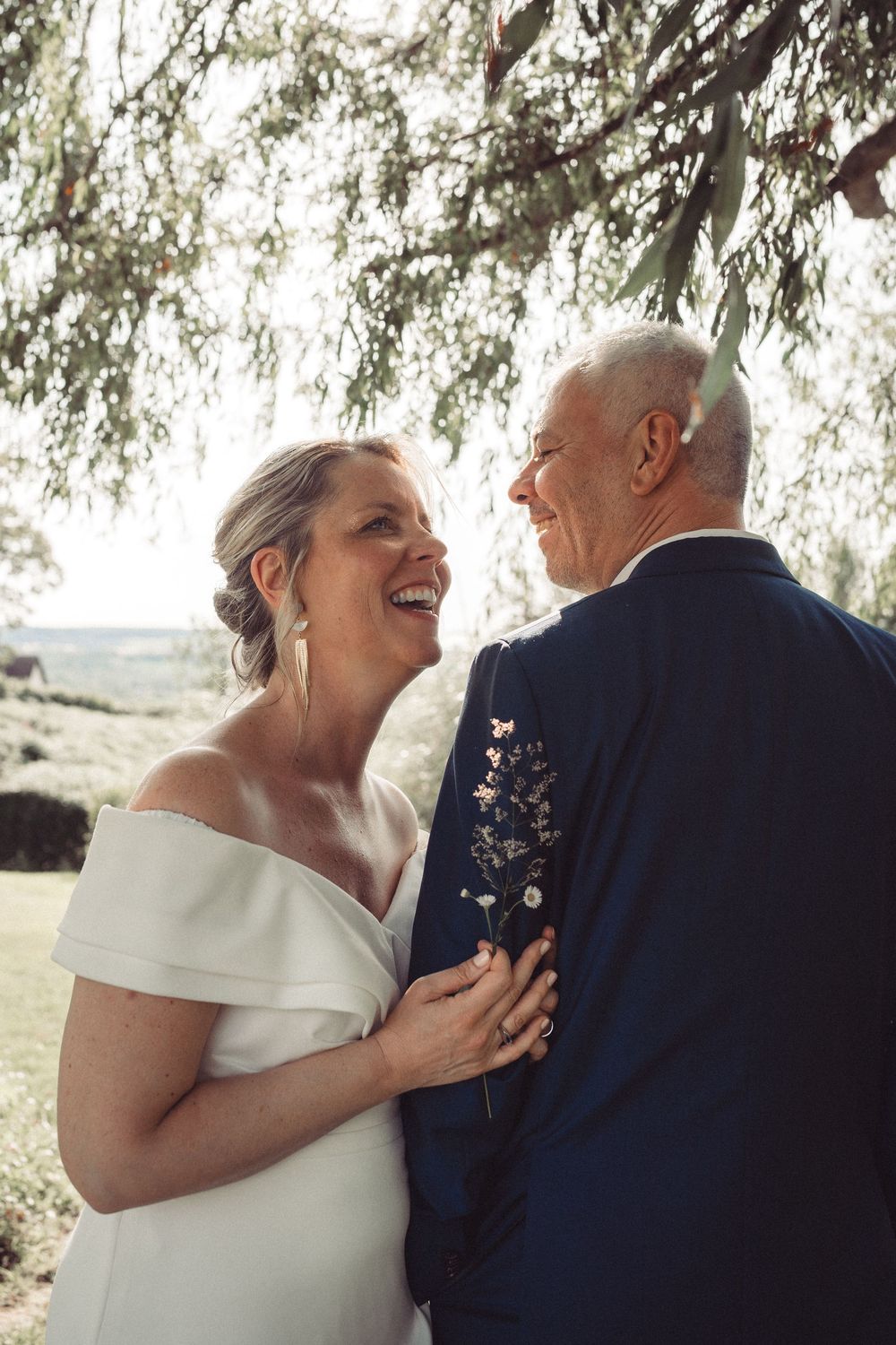 A joyful bride in an off-shoulder white gown laughs with an older groom in a navy suit under a willow tree.