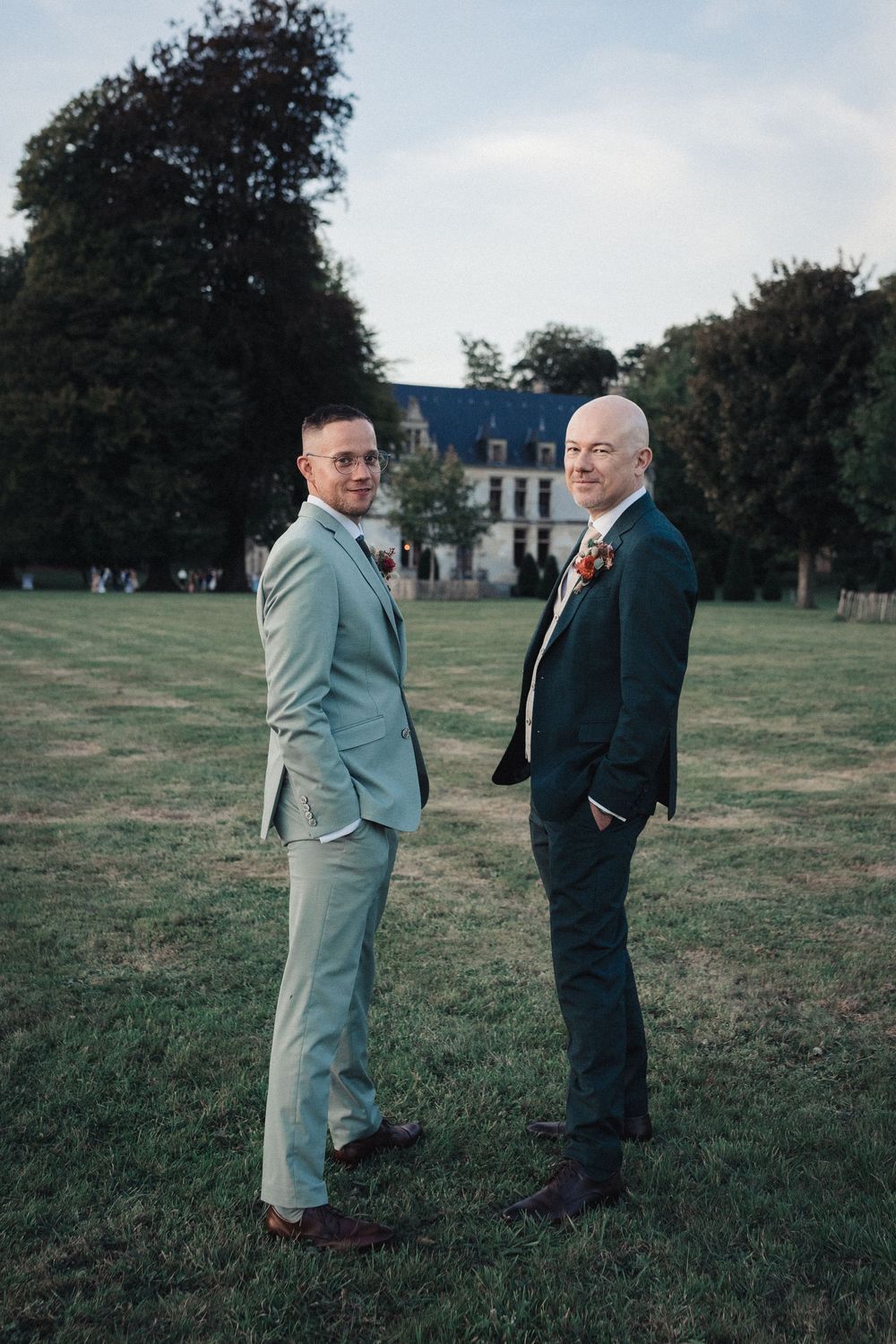 Two men in contrasting suits having a conversation on a grassy field with trees in the background.