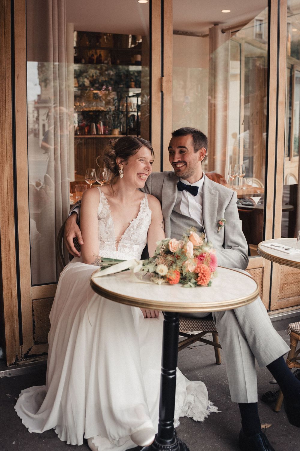 A couple shares joyful moments at their wedding reception, sitting at bistro tables in an elegant indoor venue.