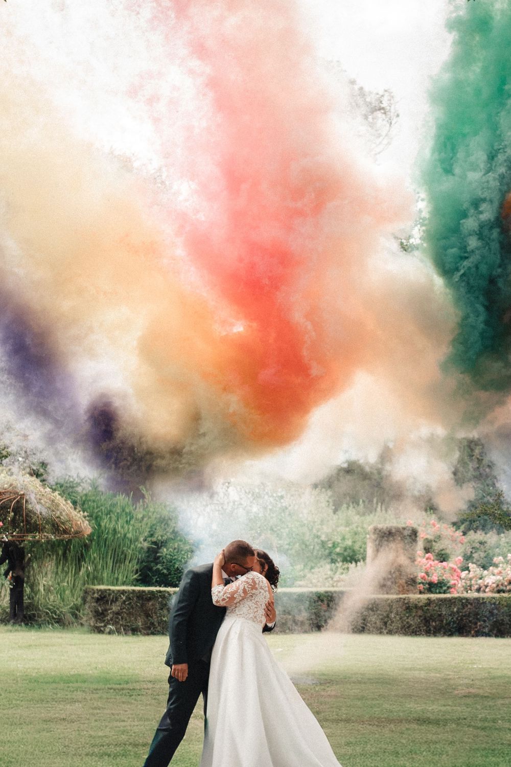 Bride and groom kissing surrounded by vibrant rainbow colored smoke bombs in a lush garden during their wedding celebration.
