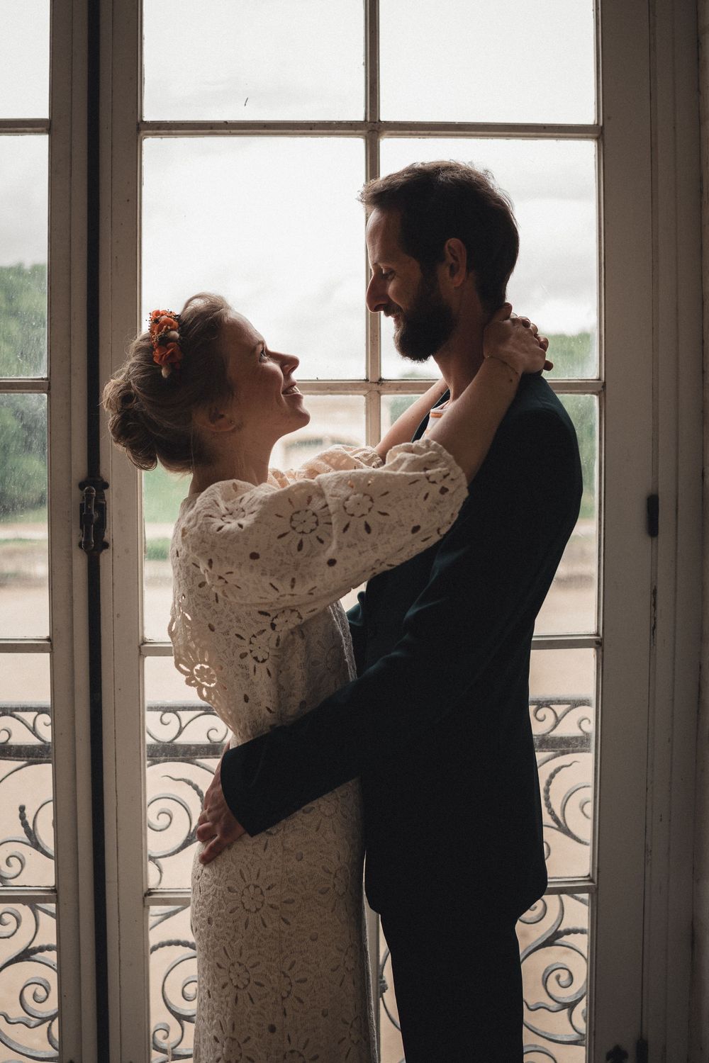 A bride and groom embrace tenderly by an ornate French window, sharing a romantic moment on their wedding day.