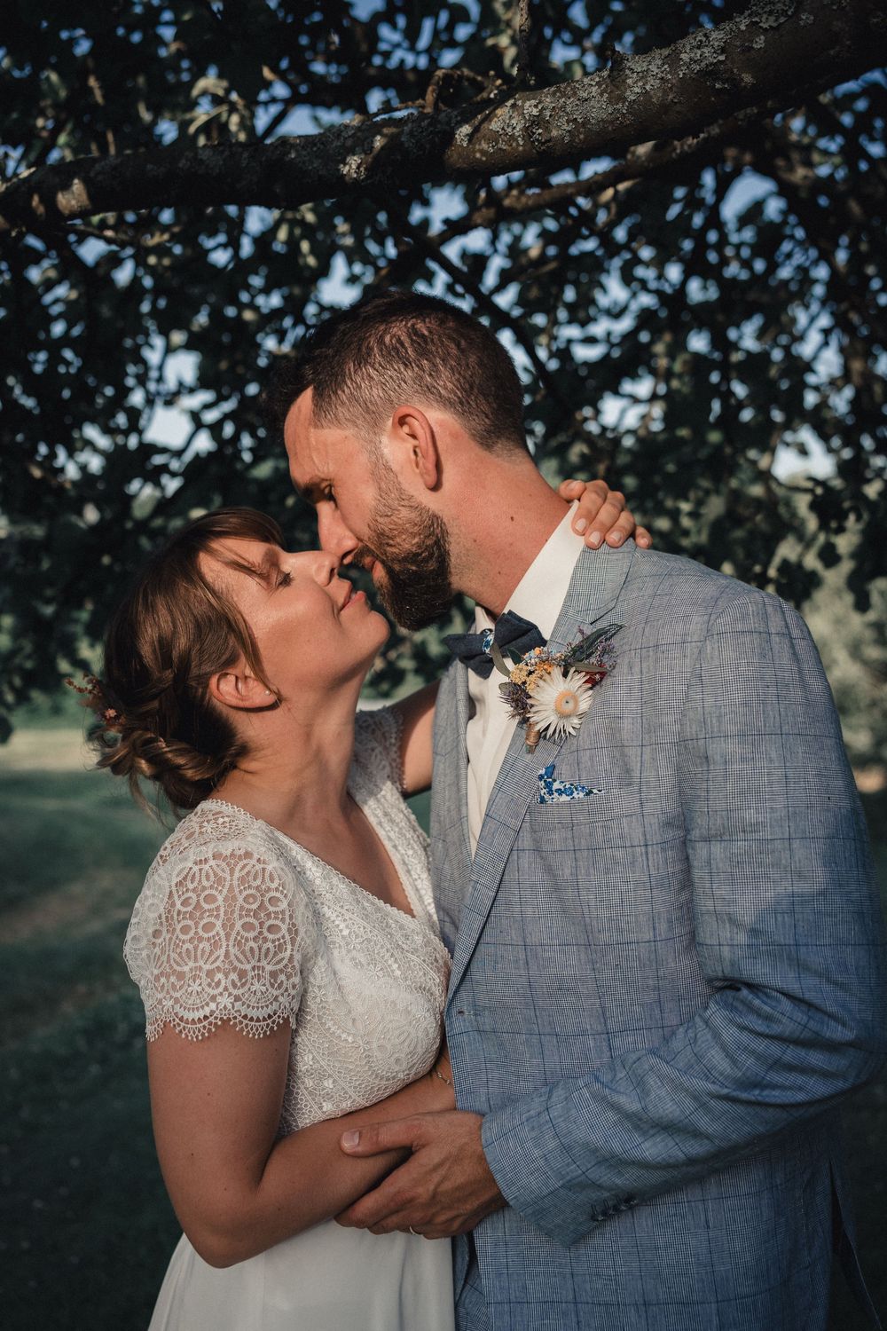 A romantic couple shares a tender kiss outdoors with trees in the background, wearing grey suit and lace dress.