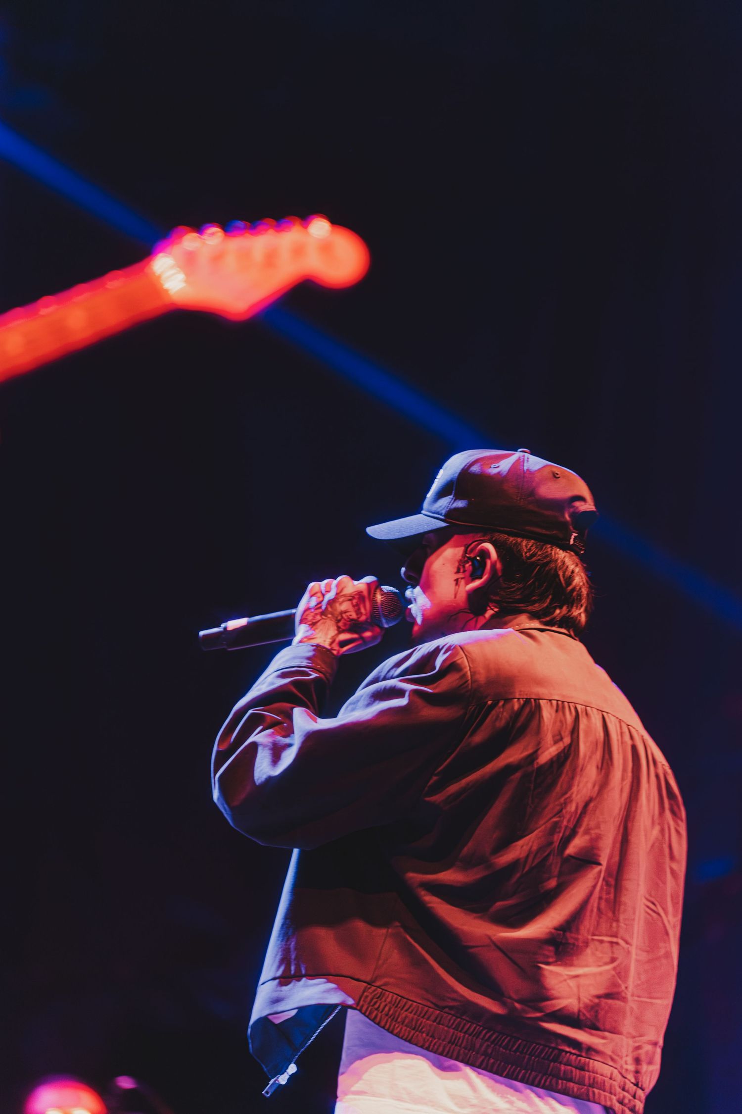 A performer in a baseball cap sings on stage under dramatic red and blue lighting during a concert performance.