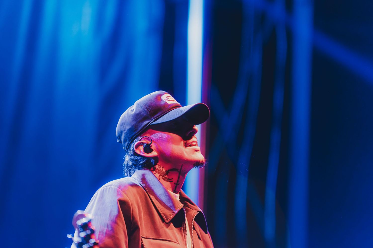A performer in a red outfit and baseball cap sings passionately on stage with dramatic blue lighting in the background.