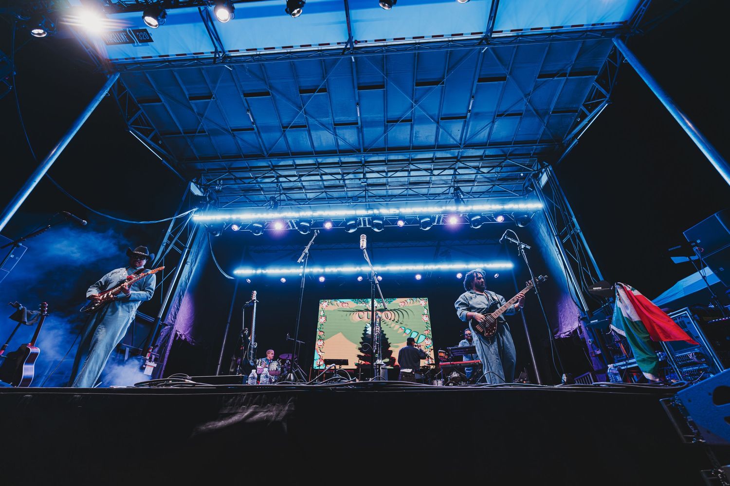 Musicians perform on an outdoor stage at night illuminated by blue lighting and spotlights.