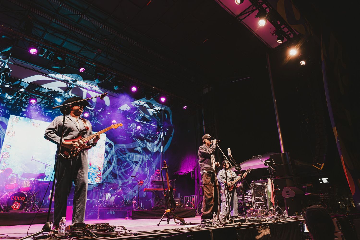A band performs on stage with colorful purple lighting and visual effects in the background during a live concert.