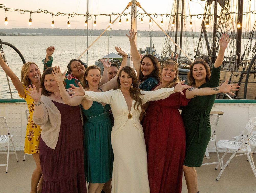 Group of people celebrating with raised arms on a dock with boats and marina in the background during sunset.