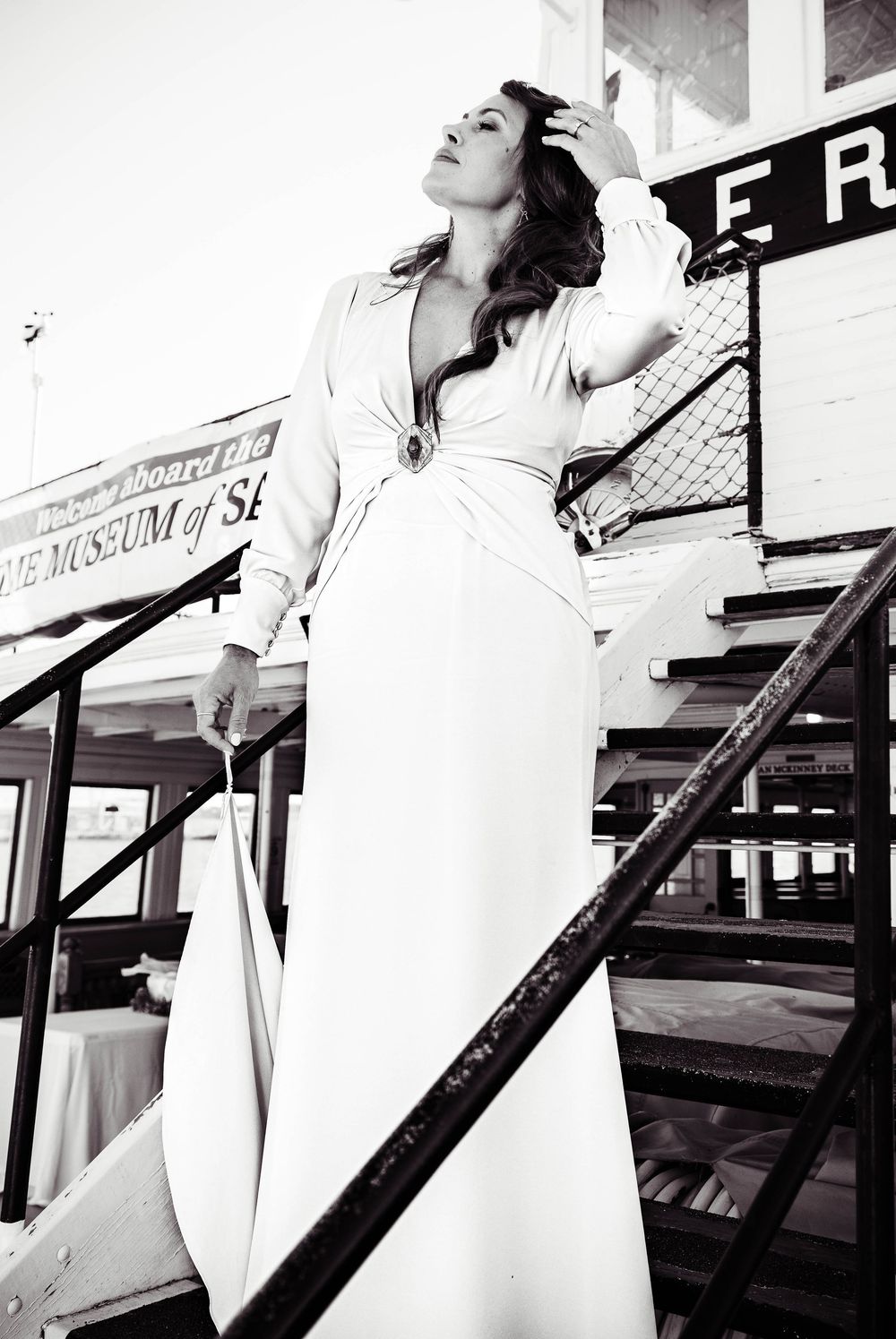 A black and white fashion photograph shows a woman in an elegant white dress posing dramatically on outdoor stairs.