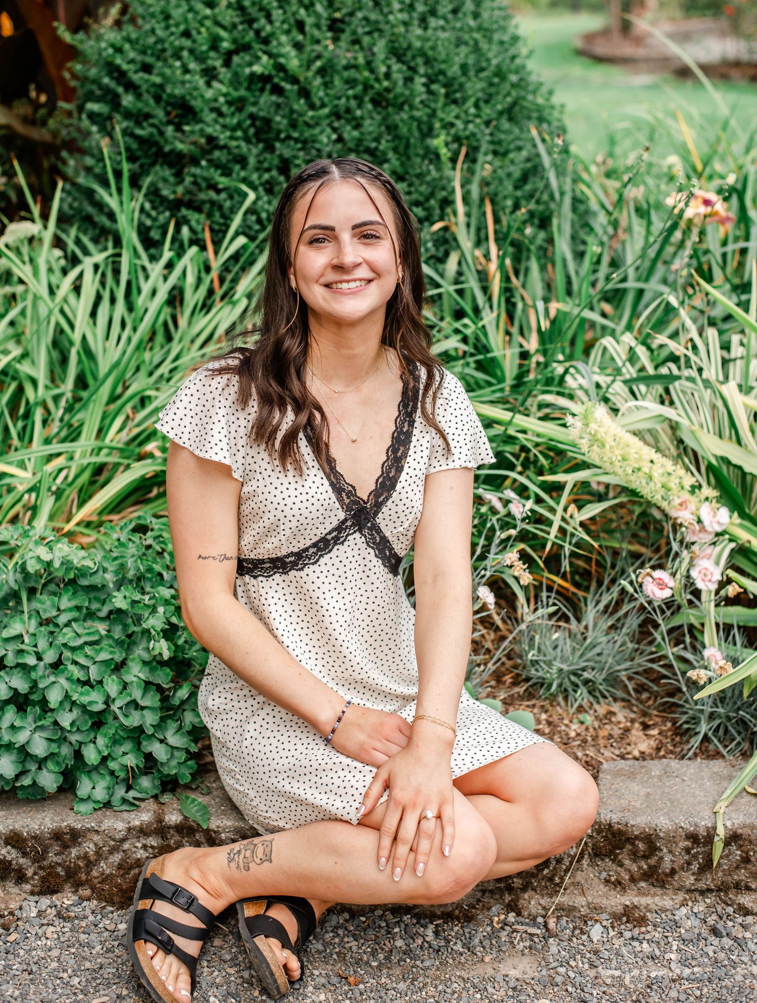 A person wearing a polka dot dress sits casually on a garden path surrounded by lush greenery and plants.