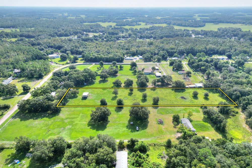 Aerial view of a lush green rural property with scattered trees, open fields, and surrounding woodland in a suburban countryside setting.