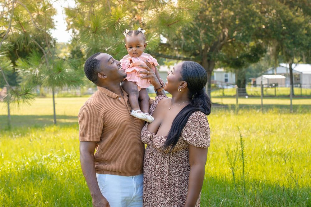 A joyful Black family with parents holding their baby girl in a sunny green park, smiling and bonding outdoors.