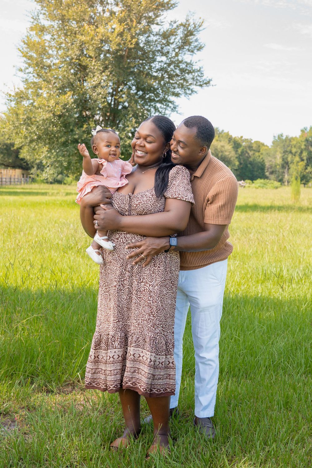 Happy Black family posing outdoors, mother holding baby girl, father embracing them in a lush green field.