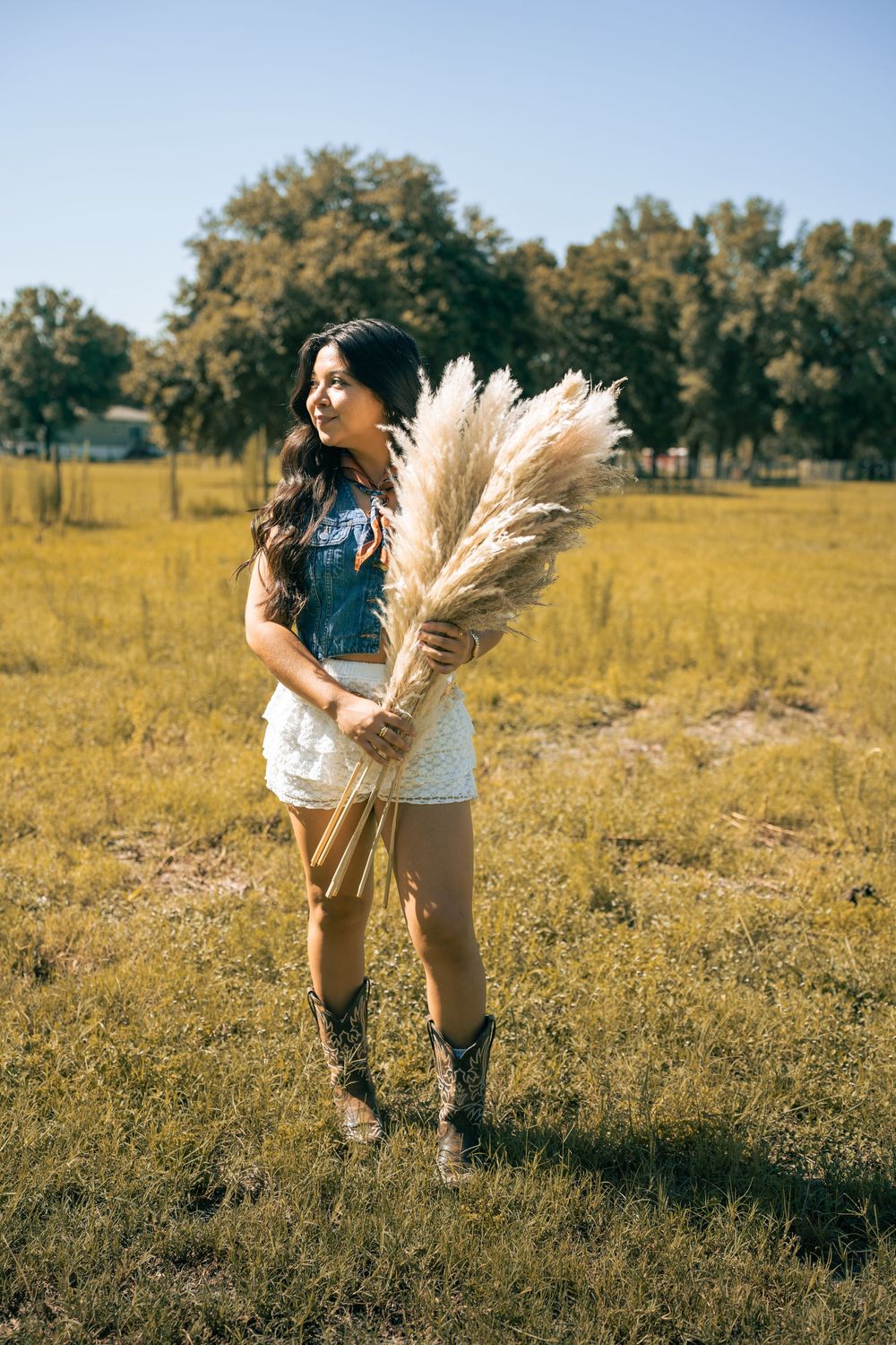 Young woman in denim vest, white skirt, and cowboy boots holding pampas grass in a sunny green field.