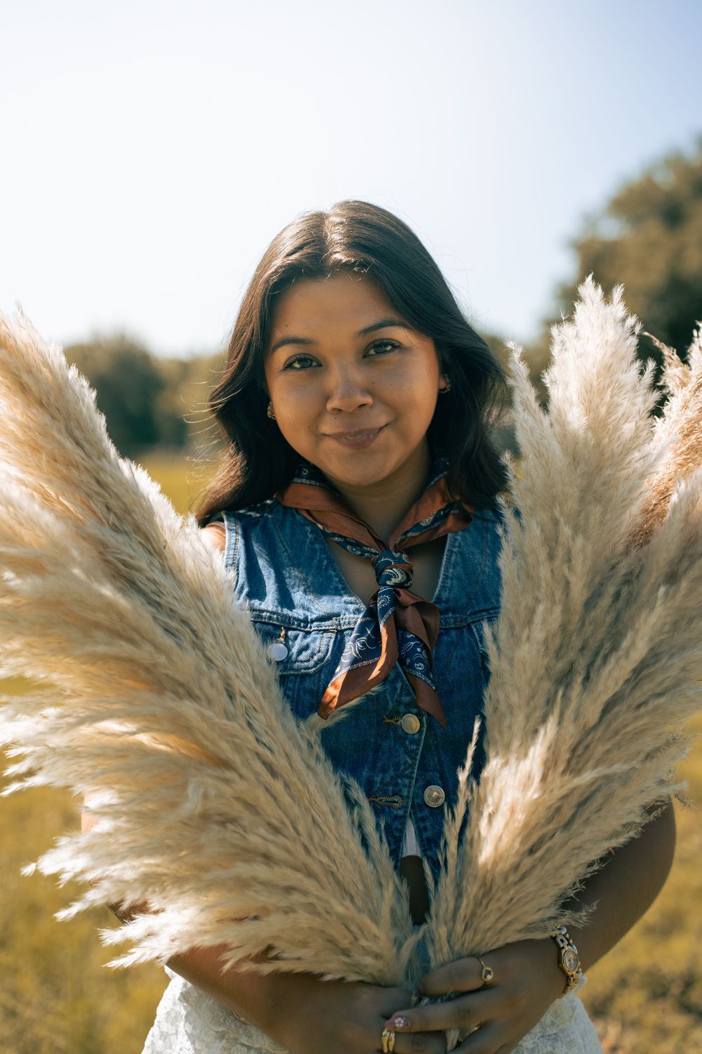 Young woman in denim vest holding pampas grass bouquet outdoors, smiling in a sunlit field.