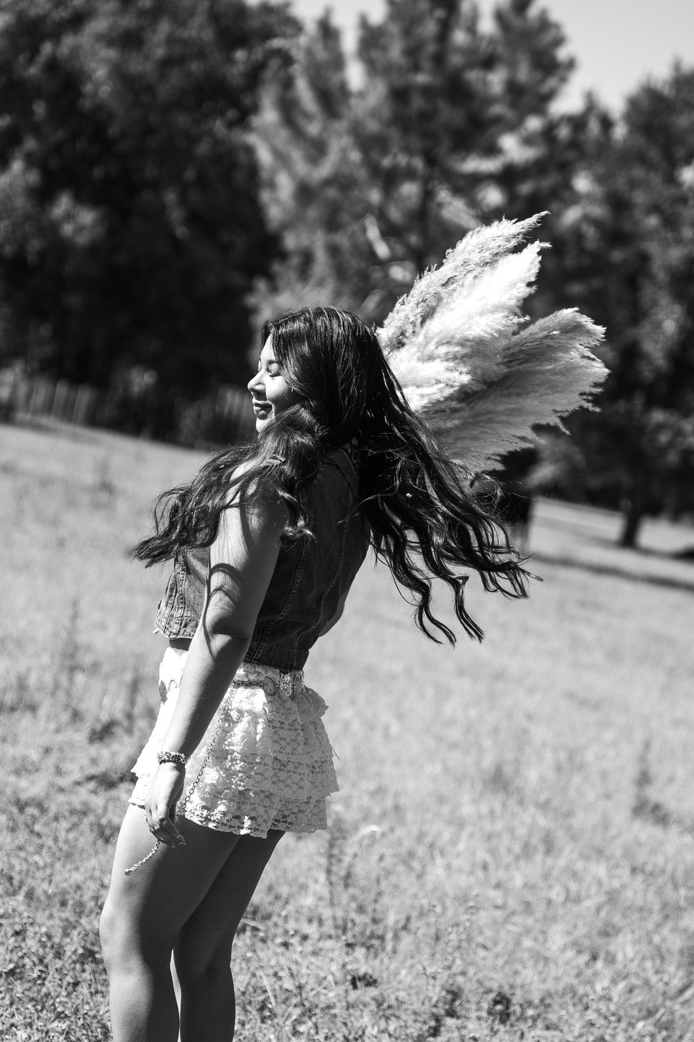 Black and white photo of a smiling woman with angel wings and sequin skirt twirling in a sunny outdoor field.