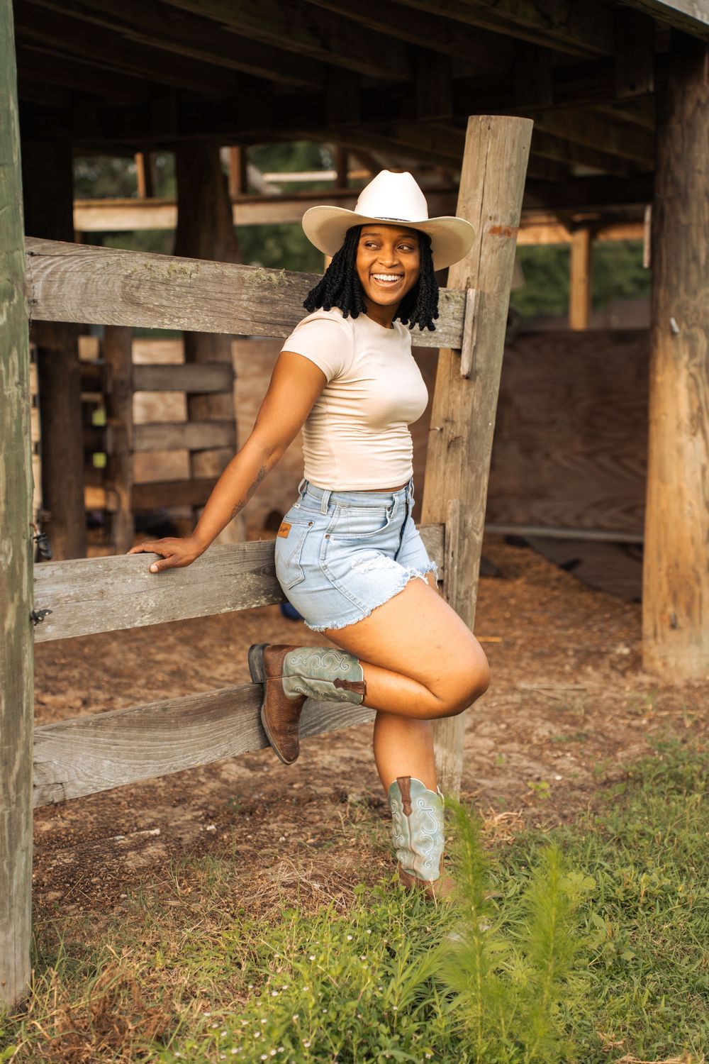 A smiling woman in a white cowboy hat, denim shorts, and cowboy boots poses against a wooden fence on a farm.