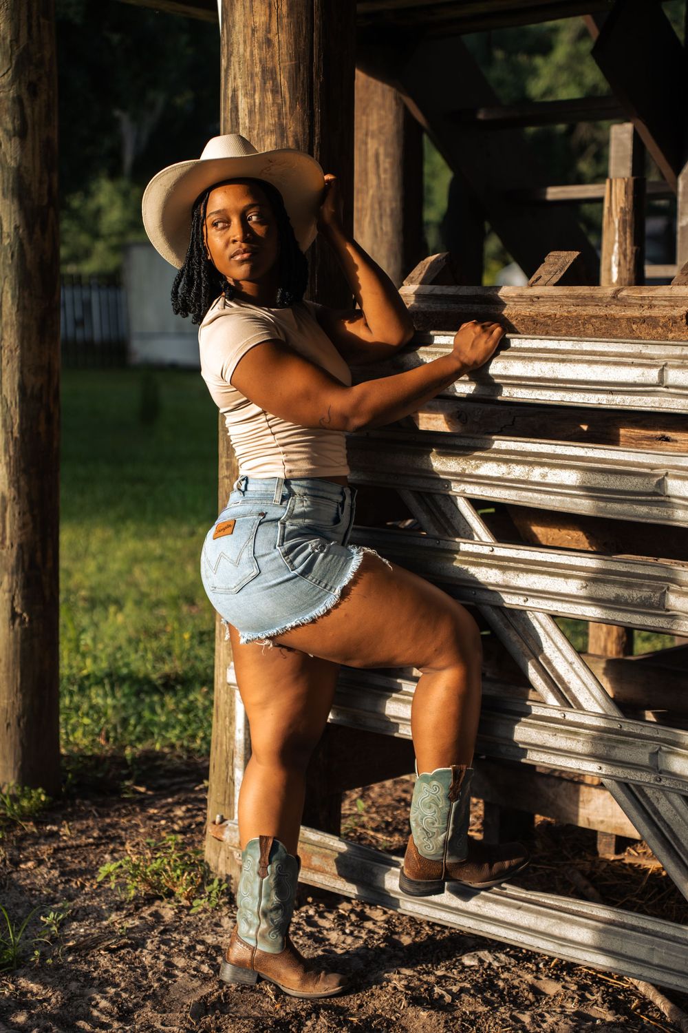 A young Black woman in cowboy hat, denim shorts, and boots poses against rustic wooden fence in golden hour light.
