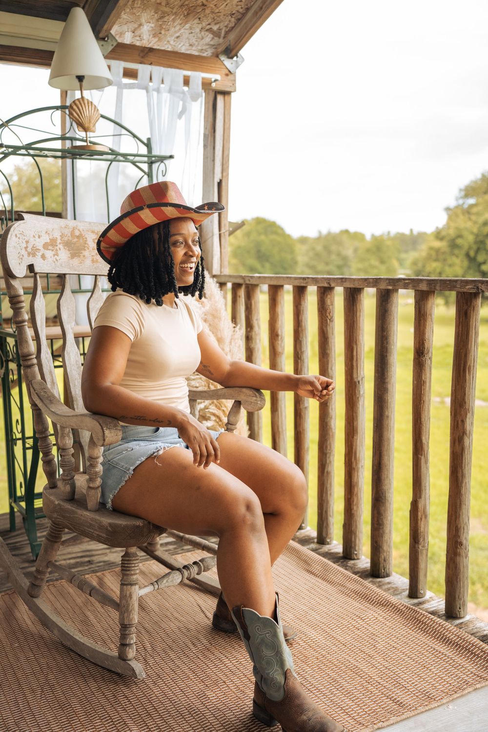 A young Black woman in a cowboy hat, denim shorts, and boots relaxes in a rocking chair on a rustic wooden porch.