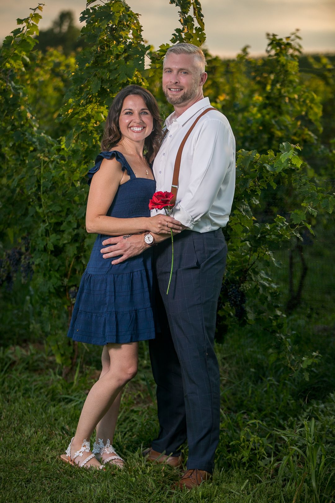 Romantic couple poses together in vineyard at sunset wearing navy dress and white shirt with suspenders.