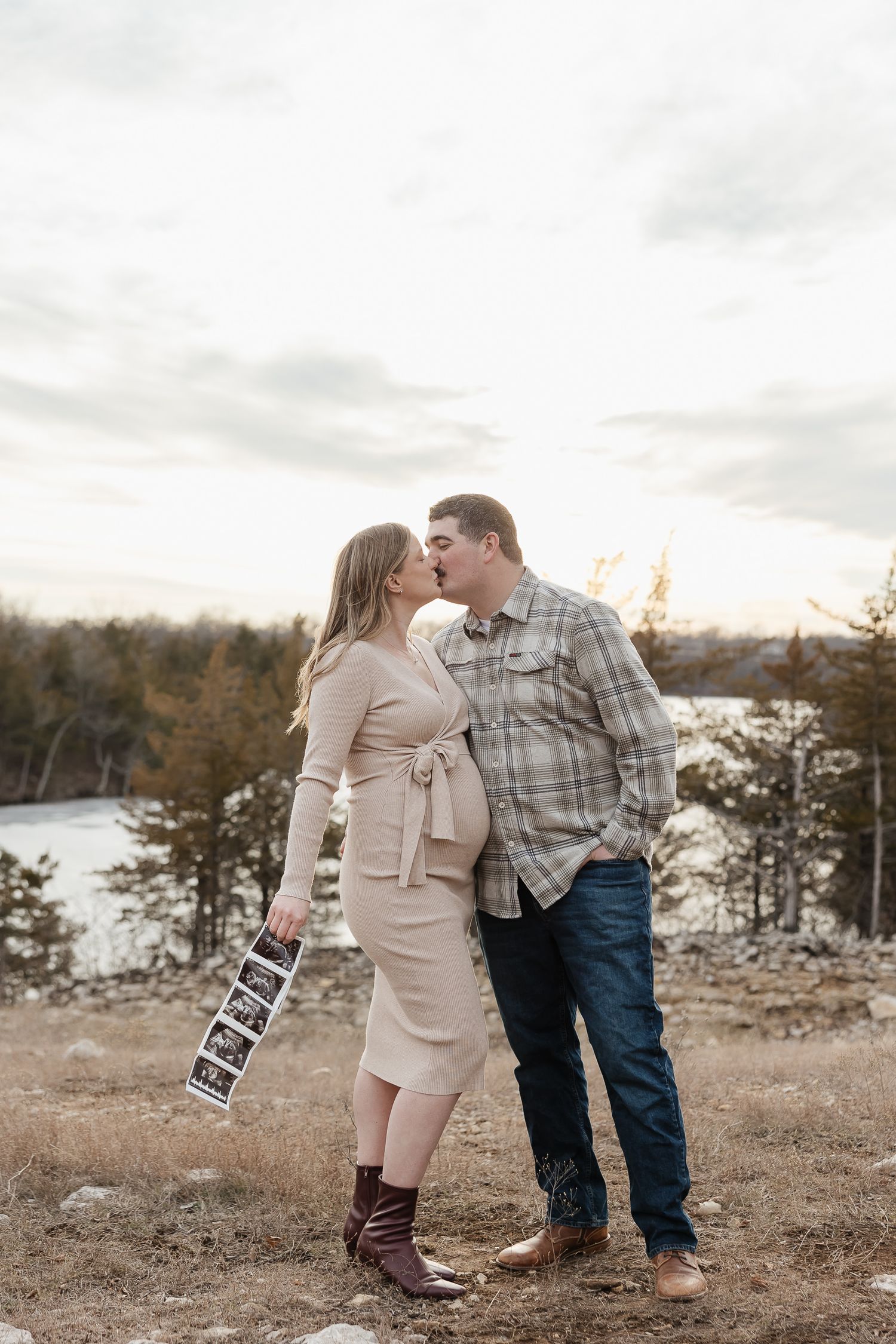 pregnant mother holds sonogram while giving kiss to and dad to be in front of lake olathe during photo session