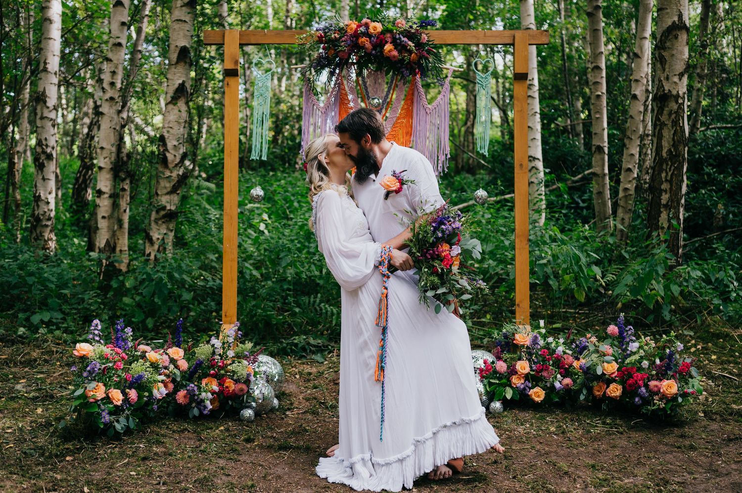 Couple kissing under a wooden arch adorned with colorful flowers in a forest wedding ceremony.